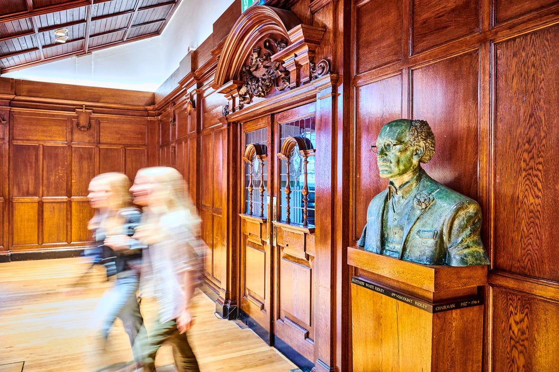 Students walking in the Armstrong building, blurred to infer movement, next to a bust