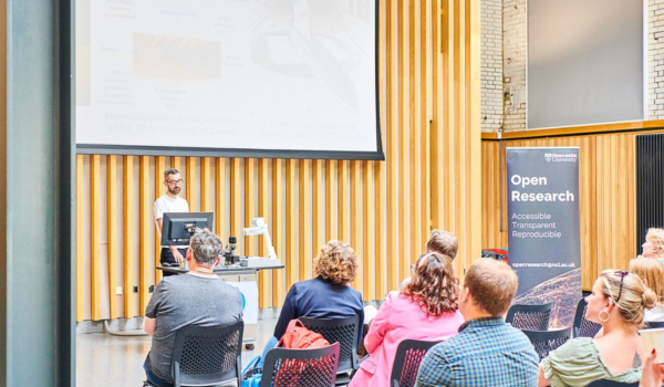 Audience members listening to a speaker presenting at a university open research event in a modern lecture space, the Boiler House