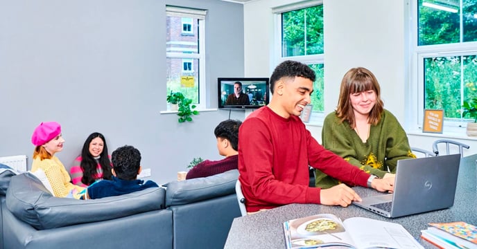 Four student sit on a couch talking between themselves, while two students sit at a table using a laptop. It's a sunny environment and a television is playing in the corner of the room. Open books are splayed out on the table in front of the two students with the laptop.
