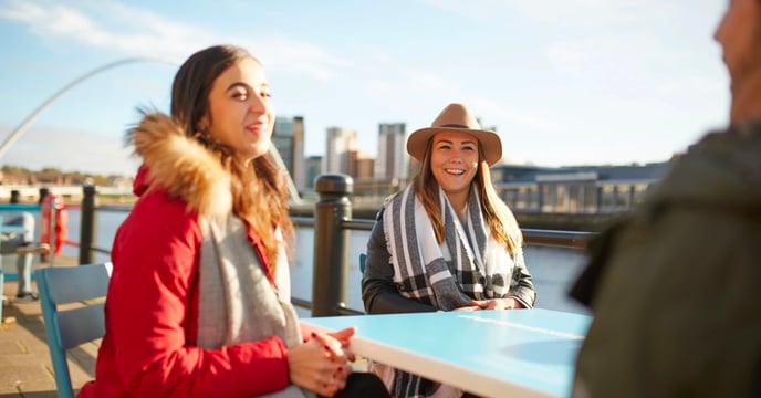Students sit at tables on the Quayside in Newcastle - the River Tyne with the Millennium Bridge can be seen in the background.