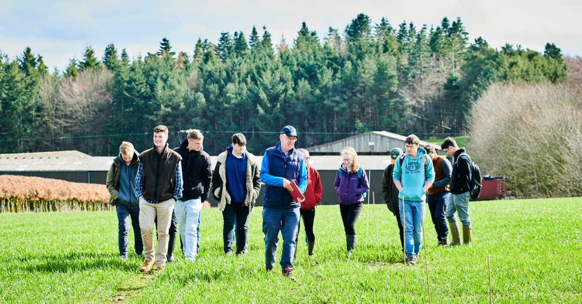 A crowd of students in cold weather clothing follow an instructor through a green field, with evergreen trees and farm buildings in the background.