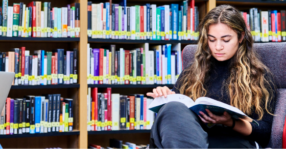 Student sat reading book on lap in front of library book shelves