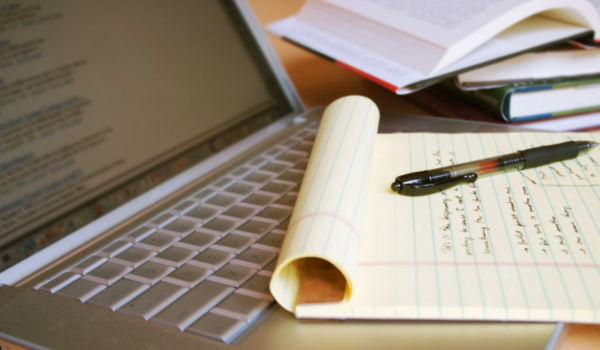 Laptop keyboard beside an open notebook with handwritten notes and a pen, with books in the background.