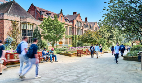 Students walking through the old quadrangle on campus with red-brick buildings, trees and outdoor seating on a sunny day.