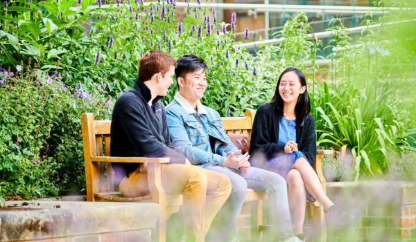 Three international students on campus sitting on a bench in summer