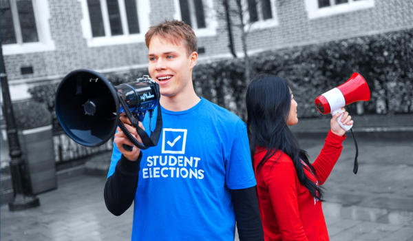 Two students stand back to back outdoors holding megaphones. One wears a blue “Student Elections” T-shirt and speaks into a black megaphone; the other wears a red top and holds a red and white megaphone, with a university building in