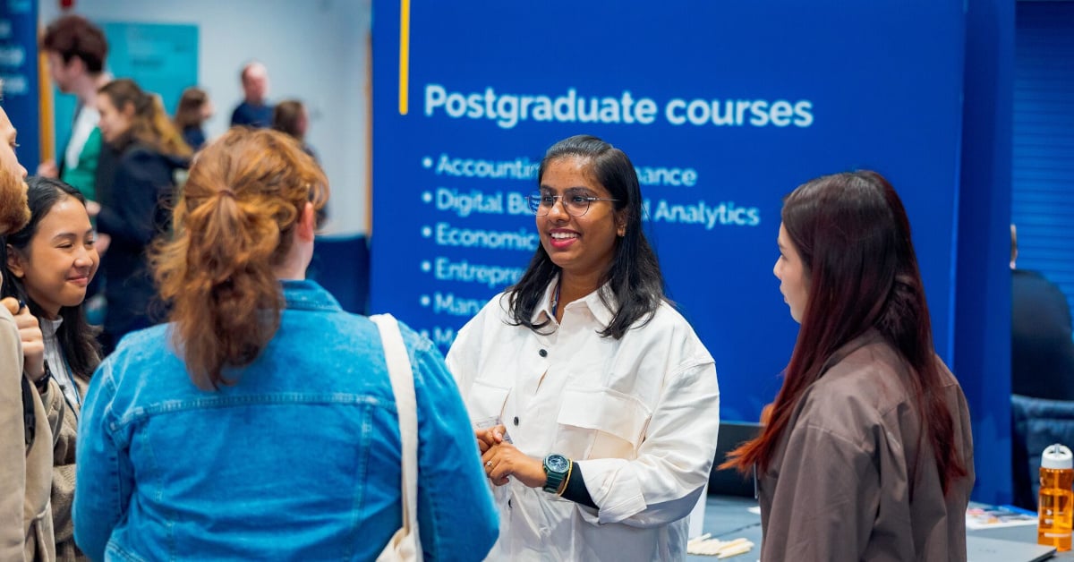 A group of postgraduate students surround a speaker during a university Open Day. In the background, is a poster, reading: 'Postgraduate courses, Accounting and finance, Economics, Entrepreneurs', and several others that are illegible.