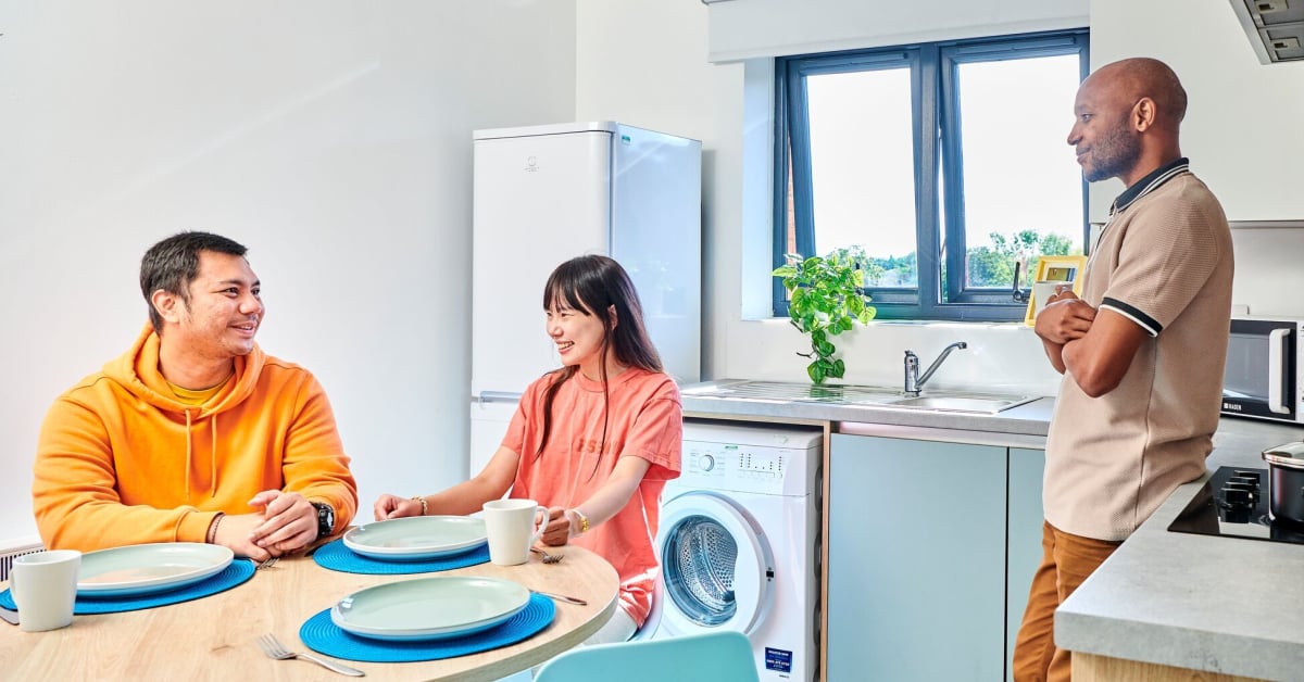 Three students hang out in a student flat, with two sitting at a table and one standing and leaning against a kitchen bench.