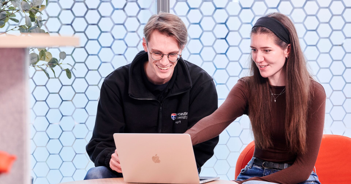 Two students smile while looking and pointing at a laptop screen.