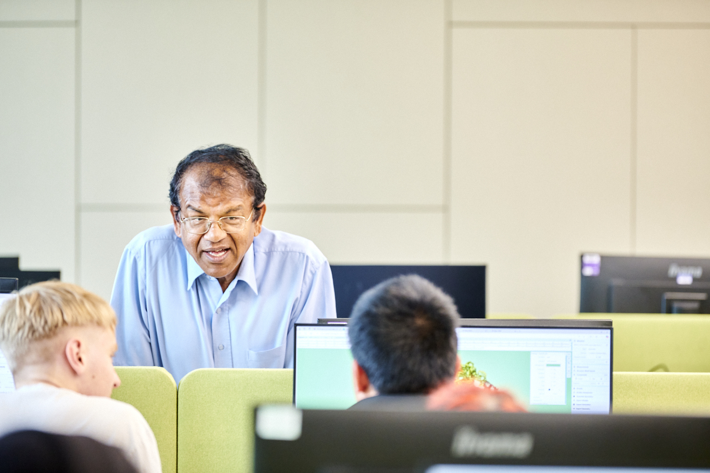 An academic leans over the back of some computers, interacting with and helping students with their work.